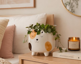 May include: A white ceramic cat-shaped planter with painted orange and brown spots, closed eyes, and a small nose. The planter features four short legs and contains a trailing green plant with small heart-shaped leaves, resting on a wooden surface.