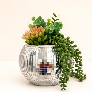 May include: A spherical, silver disco ball planter filled with assorted faux succulents. The planter has a mosaic tile design and contains green, orange, and yellow artificial plants. The overall aesthetic is modern and decorative.