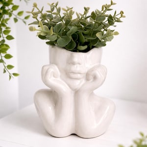 May include: A white ceramic face planter with a thoughtful pose, holding its hands up to its face. The planter is filled with green leafy faux plants. The planter is sitting on a white shelf.