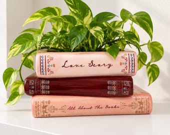May include: A ceramic planter shaped like a stack of three books with a green pothos plant. The top book is light pink with the text Love Story, the middle is dark red, and the bottom is light pink with the text All About The Books.