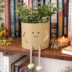 May include: A whimsical, speckled beige ceramic planter with a smiling face, arms, and dangling legs. The planter holds green foliage and sits on a wooden shelf with books and a lit candle. A charming decorative item.
