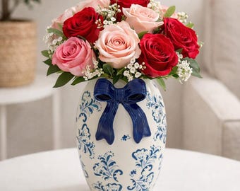 May include: A bouquet of red and pink roses with white filler flowers in a white vase. The vase has a blue bow and blue floral patterns. The arrangement is on a white table.