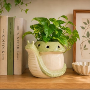 May include: A whimsical, snail-shaped ceramic planter in cream and light green, filled with vibrant green pothos plant. The planter has a detailed design, including a snail's body and head. The plant pot is on a wooden shelf with books and a framed botanical print.