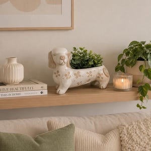May include: A cream-colored ceramic dachshund-shaped planter featuring a repeating pattern of small, light brown flower motifs across its body. The dog-shaped vessel holds a small green plant and is positioned on a wooden shelf.
