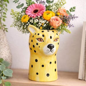 May include: A yellow ceramic vase shaped like a cheetah's head, adorned with black spots. The vase is filled with a vibrant arrangement of pink gerbera daisies, yellow ranunculus, and peach roses, creating a colorful floral display.