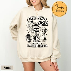 May include: A sand-colored crewneck sweatshirt featuring a black graphic of a skeleton with a messy bun and glasses, holding a drink. The text reads: I ASKED MYSELF IF I'M OKAY AND WE BOTH STARTED LAUGHING. The design includes small star accents.