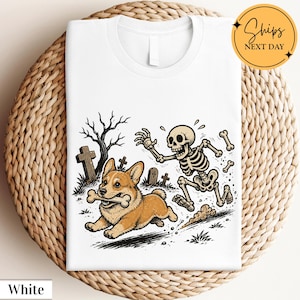 May include: A white t-shirt featuring a graphic illustration of a brown and white Corgi dog running with a bone in its mouth, being chased by a cartoon skeleton in a graveyard setting with bare trees and tombstones in the background.