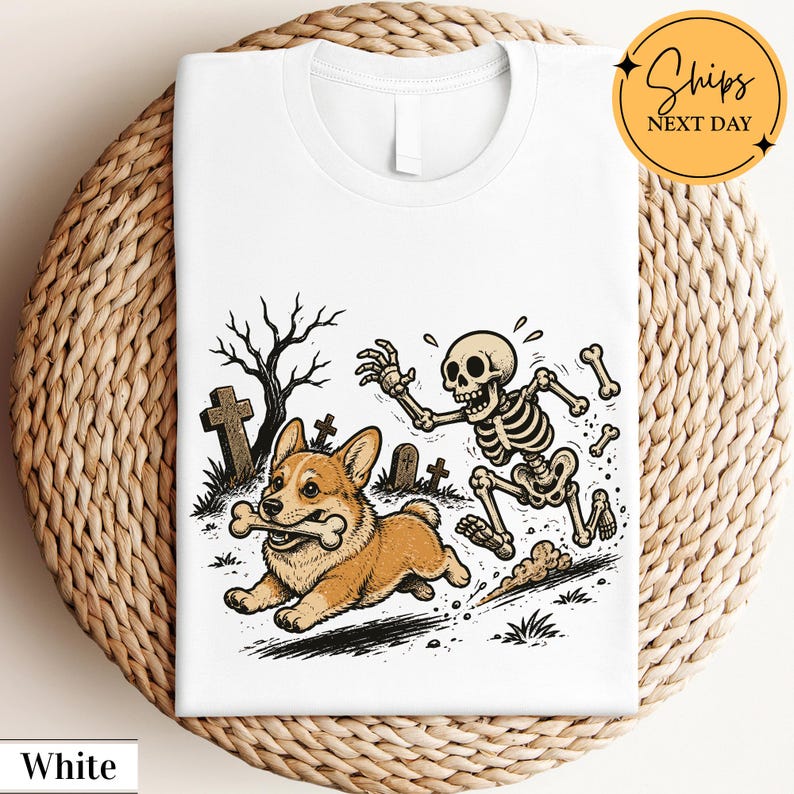 May include: A white t-shirt featuring a graphic illustration of a brown and white Corgi dog running with a bone in its mouth, being chased by a cartoon skeleton in a graveyard setting with bare trees and tombstones in the background.