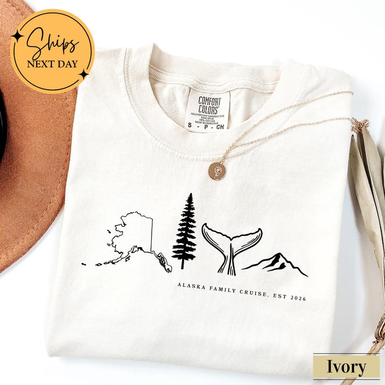 a white t-shirt with a graphic design depicting Alaska, including a tree, mountains, and a whale's tail. The text on the t-shirt reads "ALASKA FAMILY CRUISE. EST 2026".