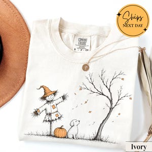 May include: An ivory t-shirt featuring a graphic illustration of a scarecrow wearing an orange hat, a small white dog, a pumpkin, and a bare tree with falling leaves. The shirt has a crew neckline and a Comfort Colors brand label at the collar.