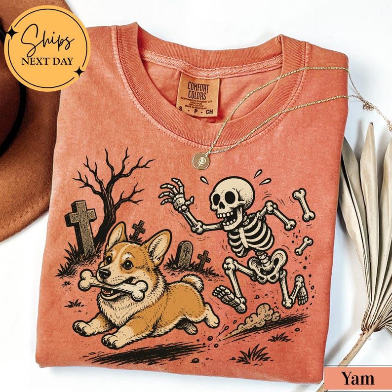 May include: A terracotta-colored t-shirt featuring a graphic print of a corgi running with a bone in its mouth while being chased by a skeleton in a graveyard setting with tombstones and a bare tree. The shirt has a Comfort Colors brand label.