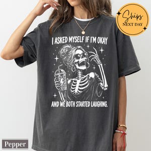 May include: A charcoal gray short-sleeve t-shirt featuring a white graphic of a skeleton wearing sunglasses and a messy bun, holding a drink. The text reads I ASKED MYSELF IF I'M OKAY AND WE BOTH STARTED LAUGHING. The shirt has a relaxed fit.