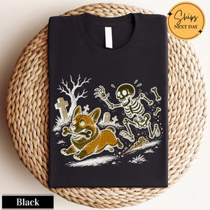 May include: A black t-shirt featuring a graphic illustration of a brown corgi running with a bone in its mouth, pursued by a white skeleton in a graveyard setting with tombstones and a bare tree. The design is rendered in a detailed line art style.