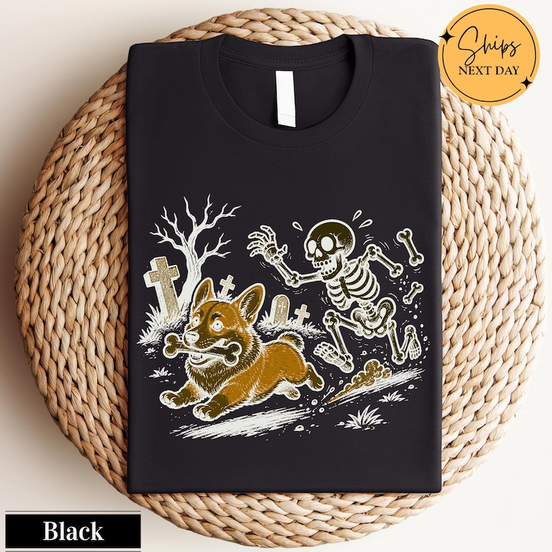 May include: A black t-shirt featuring a graphic illustration of a brown corgi running with a bone in its mouth, pursued by a white skeleton in a graveyard setting with tombstones and a bare tree. The design is rendered in a detailed line art style.