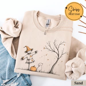 May include: A sand-colored crewneck sweatshirt featuring a graphic illustration of a scarecrow, a small dog, a pumpkin, and a bare tree with falling leaves. The design is rendered in black ink with orange accents on the hat and pumpkin.