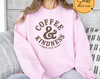 May include: Light pink sweatshirt with the text "Coffee & Kindness" in brown lettering. The sweatshirt also has the phrase "A Brew-tiful Blend" printed below the main text. The image also includes a "Ships Next Day" graphic.