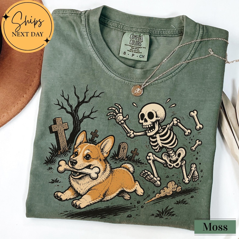 May include: A moss green t-shirt featuring a graphic print of a Corgi dog running with a bone in its mouth while being chased by a cartoon skeleton in a graveyard setting with tombstones and a bare tree. The shirt has a crew neck and a tag.