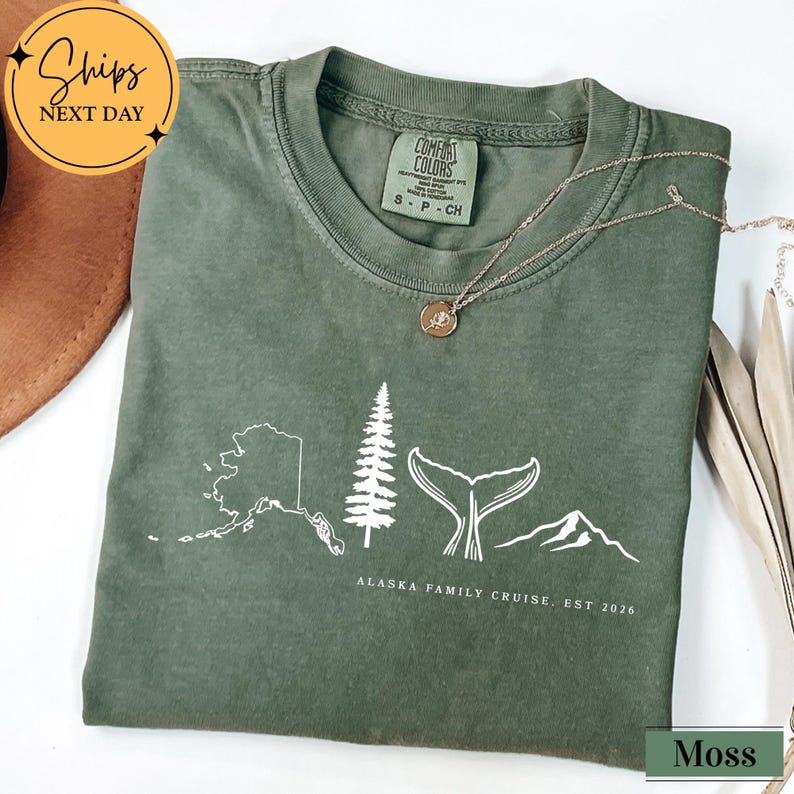 A green t-shirt with a graphic design depicting a tree, mountains, and a whale, along with text that says "Alaska Family Cruise, est 2026".