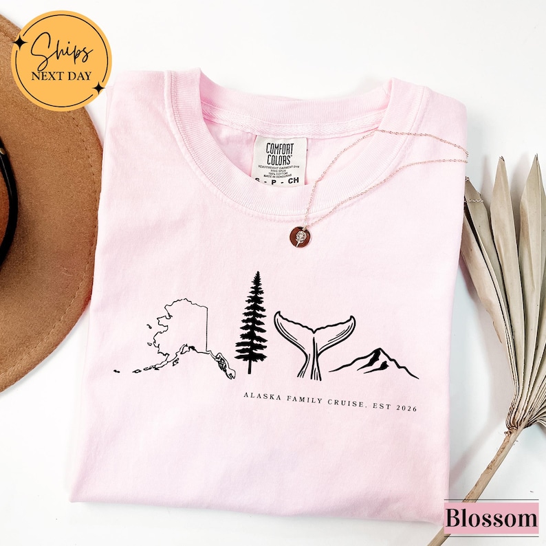 A pink t-shirt with a graphic design depicting Alaska, including a tree, mountains, and a whale, along with the text "ALASKA FAMILY CRUISE, EST 2016".