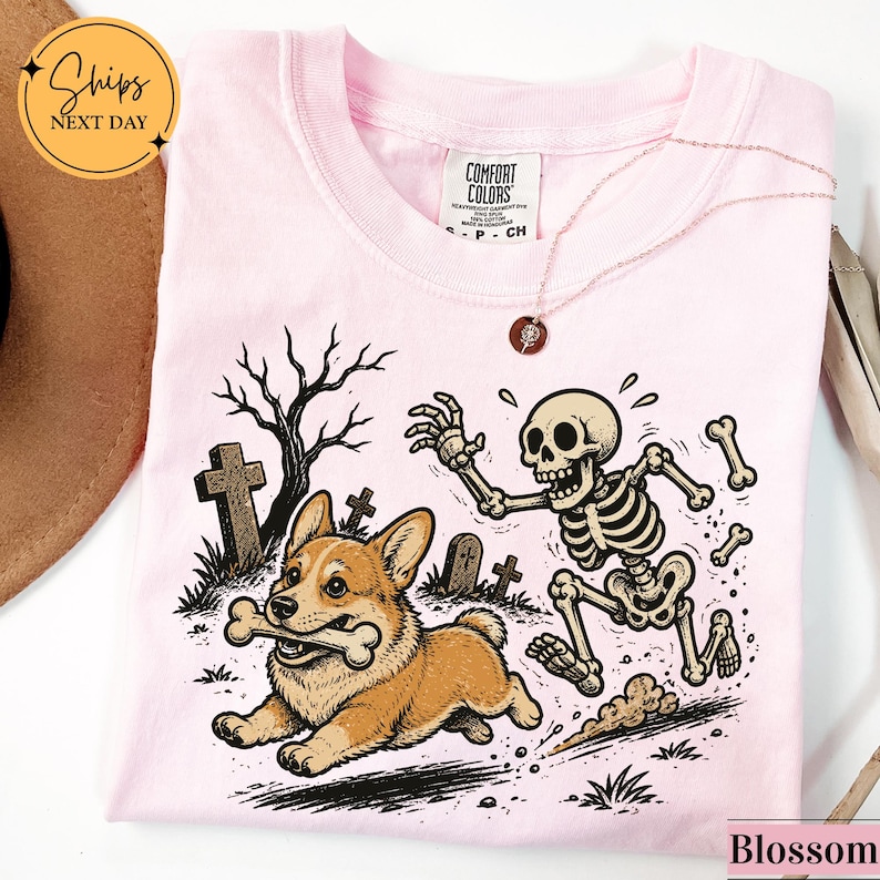 May include: A light pink t-shirt featuring a graphic print of a Corgi dog running with a bone in its mouth while being chased by a cartoon skeleton in a graveyard setting with tombstones and a bare tree. The shirt has a Comfort Colors brand label.