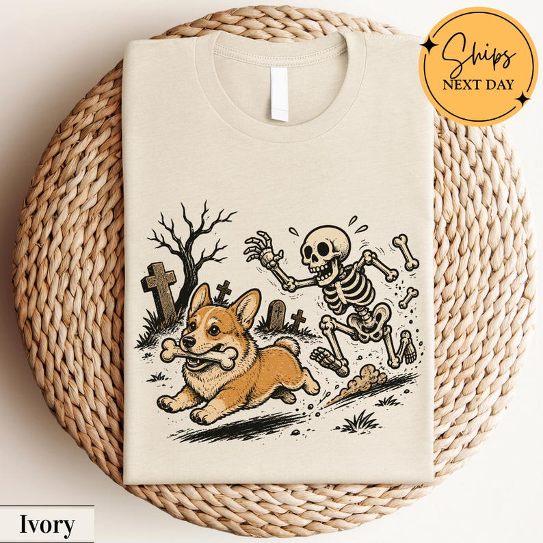 May include: An ivory t-shirt featuring a graphic illustration of a brown and white Corgi dog running with a bone in its mouth, pursued by a cartoon skeleton in a graveyard setting with bare trees and tombstones under a light background.