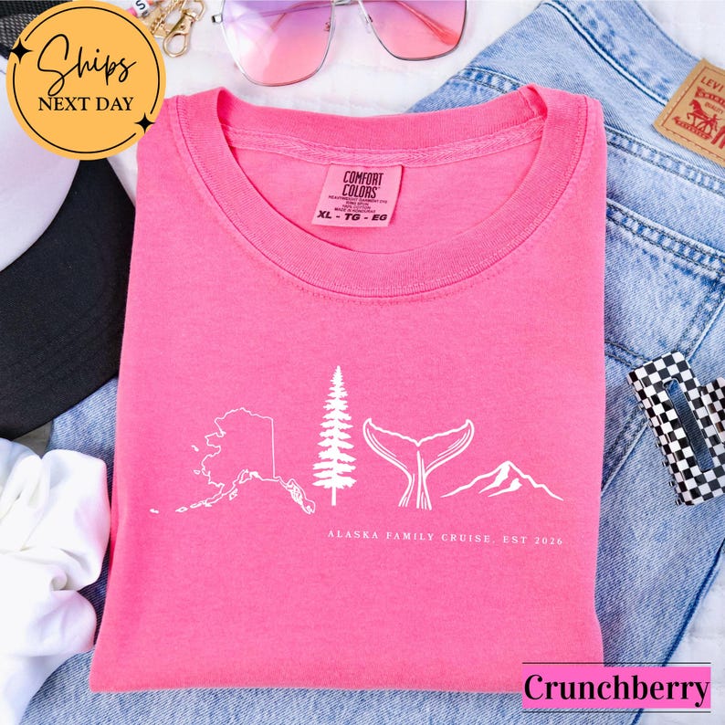 A pink t-shirt with a graphic design of Alaska, a family cruise, and mountains, along with the text "Alaska Family Cruise. Est 2020" printed on it. The t-shirt is folded and placed on top of a pair of jeans, with a pair of sunglasses and a hat visible in the background.