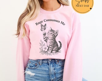 May include: Light pink sweatshirt with the text "Rage Consumes Me" above a graphic of a kitten reaching for a butterfly. The sweatshirt is a light pink color and is paired with blue jeans.