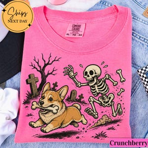 May include: A bright pink t-shirt featuring a graphic print of a brown and white Corgi dog running with a bone in its mouth, pursued by a cartoon skeleton in a graveyard setting with tombstones and a bare tree, with a Comfort Colors label.