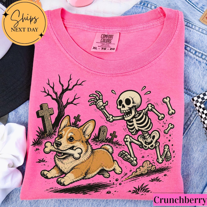 May include: A bright pink t-shirt featuring a graphic print of a brown and white Corgi dog running with a bone in its mouth, pursued by a cartoon skeleton in a graveyard setting with tombstones and a bare tree, with a Comfort Colors label.