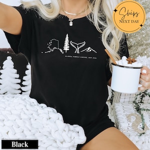 A person wearing a black t-shirt with a graphic design of Alaska, including a whale, mountain, and pine tree, holding a mug of hot chocolate.