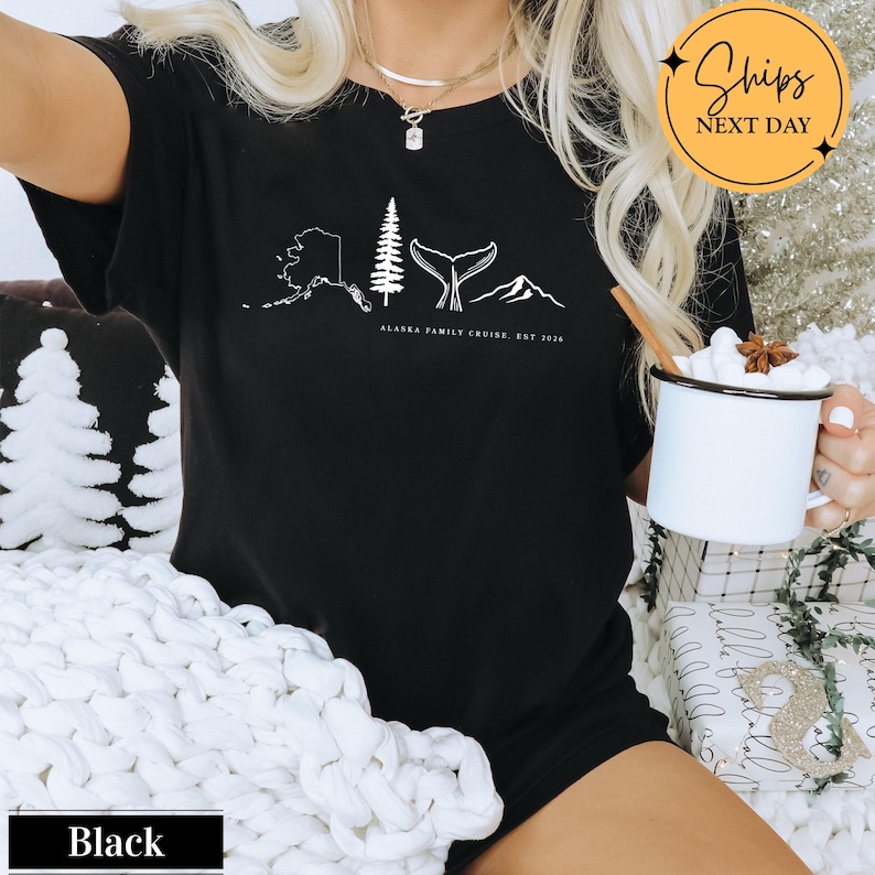 A person wearing a black t-shirt with a graphic design of Alaska, including a whale, mountain, and pine tree, holding a mug of hot chocolate.