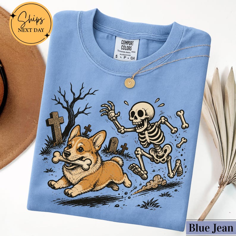 May include: A light blue t-shirt featuring a graphic of a Corgi dog running with a bone in its mouth, chased by a skeleton in a graveyard setting with tombstones and a bare tree. The shirt has a crew neckline and a Comfort Colors brand label.