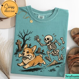 May include: A seafoam green crewneck t-shirt featuring a graphic print of a corgi running with a bone in its mouth while being chased by a skeleton in a graveyard setting with tombstones and a bare tree. The shirt has a Comfort Colors label.