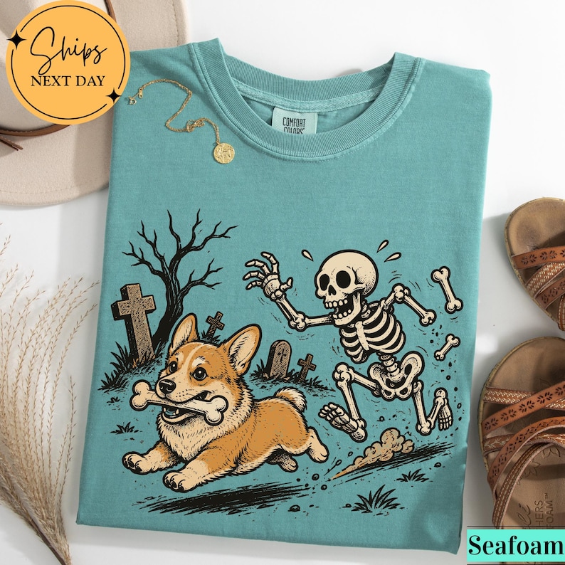 May include: A seafoam green crewneck t-shirt featuring a graphic print of a corgi running with a bone in its mouth while being chased by a skeleton in a graveyard setting with tombstones and a bare tree. The shirt has a Comfort Colors label.