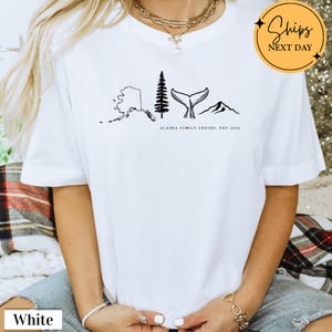 A white t-shirt with a graphic design depicting Alaska, including a tree, mountains, and a whale.