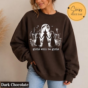 May include: A dark chocolate brown crewneck sweatshirt featuring a white graphic print of three figures holding hands in front of a large full moon and a bonfire. The text "girls will be girls" is printed in a typewriter font below the illustration.