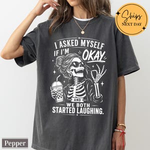 May include: A charcoal gray t-shirt featuring a white graphic of a skeleton wearing sunglasses, holding a drink, and making an okay hand gesture. The text reads: I ASKED MYSELF IF I'M OKAY AND WE BOTH STARTED LAUGHING. The shirt has a crew neckline.