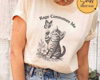 May include: Ivory-colored t-shirt featuring a black and white illustration of a kitten reaching for a butterfly. The text "Rage Consumes Me" arches above the kitten. The shirt has rolled-up sleeves and is paired with blue jeans.