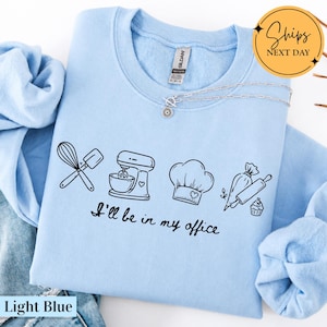 May include: A light blue crewneck sweatshirt featuring a black graphic print of a whisk, spatula, stand mixer, chef hat, piping bag, rolling pin, and cupcake. The text "I'll be in my office" is printed in black script below the baking illustrations.