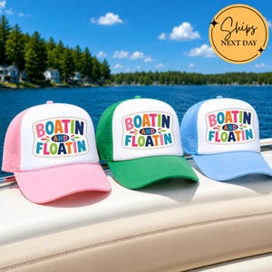 May include: Three trucker hats with white front panels and mesh backs in pink, green, and light blue. Each hat features a colorful graphic patch with the text "BOATIN AND FLOATIN" in a playful, multi-colored font with decorative accents.