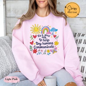 May include: Light pink sweatshirt with a colorful graphic that includes a smiling sun, rainbow, hearts, flowers, and butterflies. The text on the sweatshirt reads "it's a good day to help tiny humans communicate."
