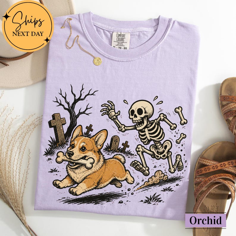 May include: A light purple t-shirt featuring a graphic illustration of a corgi running with a bone in its mouth while being chased by a skeleton in a graveyard setting with bare trees and tombstones. The shirt has a crew neckline and a tag.
