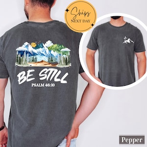 May include: Pepper-colored t-shirt with a mountain and forest graphic on the back, and a smaller mountain and cross design on the front. The back graphic includes the text "BE STILL" and "PSALM 46:10". The shirt has a crew neck and short sleeves.
