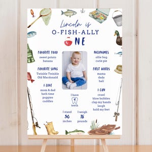 以下が含まれることがあります： 釣りテーマの1歳の誕生日記念ボード。ボート、魚、タックルボックス、網の水彩イラストが描かれています。テキストには「Lincoln is O-FISH-ALLY ONE」とあり、子供の好物やニックネーム、成長記録などの項目が記載されています。