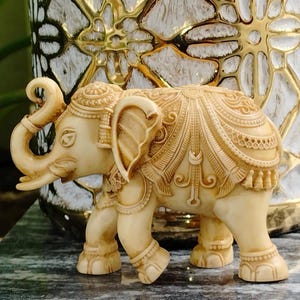 Puede incluir: Figurilla de elefante tallada en marfil, adornada con intrincados diseños en la cabeza, orejas y cuerpo, evocando un diseño tradicional indio. La figura se apoya sobre cuatro patas, con la trompa curvada hacia arriba. El fondo presenta un jarrón decorativo.