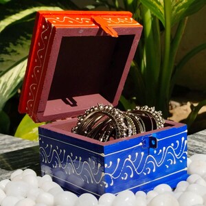 May include: A small, open wooden jewelry box with a red lid and blue base, decorated with white floral patterns. The box contains several gold bangles and sits on a bed of white pebbles. The background features lush green foliage.