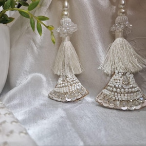 May include: Close-up of two white beaded tassels with a layered design. Each tassel features a beaded top, a white fringe, and a bottom section adorned with white beads and sequins. The tassels hang against a white, textured background.