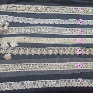 May include: Assortment of white lace trims with pearl and floral embellishments. The trims vary in design, including beaded edges, floral patterns, and delicate textures. A strand of white pearls is visible on the left side, complementing the trims.