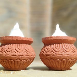 Può includere: Due piccoli vasi color terracotta con intricati disegni scolpiti. Ogni vaso è riempito con una sostanza bianca e soffice, forse una candela o un elemento decorativo. I vasi sono posti su uno sfondo sfocato.