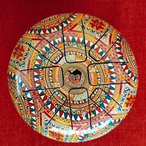 May include: A colorful steel tongue drum with a vibrant, patterned design in red, yellow, blue, and white. The drum features a central sound hole and eight cut tongues, creating a unique musical instrument.